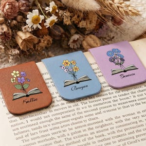 May include: Three floral magnetic bookmarks on a book page. Each bookmark features a different color: brown, blue, and lavender. Each has a floral design and a name: Kallie, Brayan, and Benicio.