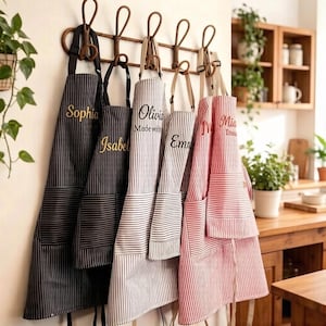 May include: Personalised aprons with vertical stripes in black, white, and red, hanging on a decorative rack. Each apron has a name embroidered in gold thread. The aprons are displayed in a kitchen setting.