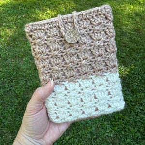 May include: A crocheted book cover in two-tone beige and cream. The top portion is a darker beige, while the bottom is cream. A wooden button and loop closure secures the cover. The cover is held against a background of green grass.