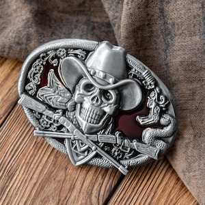 May include: Silver-coloured oval belt buckle featuring a skull wearing a cowboy hat. The buckle has a red background and is decorated with crossed rifles, a snake, and floral designs. The buckle is on a wooden surface.