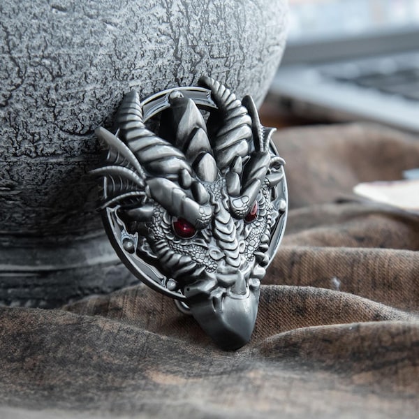 Könnte beinhalten: Silberfarbener Drachenkopf-Anhänger mit roten Augen. Der Anhänger zeigt detaillierte Schuppen und Hörner, eingefasst in einem Kreis. Der graue, strukturierte Hintergrund deutet auf ein Fantasy- oder Mittelalter-Thema hin.