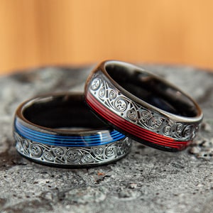 Puede incluir: Dos anillos de metal oscuro con intrincados patrones de volutas en tono plateado. Un anillo muestra una franja horizontal azul vibrante, mientras que el otro anillo presenta una franja horizontal roja llamativa en el centro del diseño detallado.