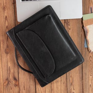 Peut inclure: Un porte-documents en cuir noir avec une poignée, une grande poche et une petite poche avec un rabat incurvé. Le porte-documents est posé sur une surface en bois à côté d'un ordinateur portable et de quelques papiers.