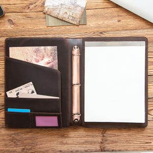 May include: Open brown leather portfolio with a notepad, card slots, and a pen holder. The portfolio is open, revealing a white notepad and several pockets. The background is a wooden surface with postcards.
