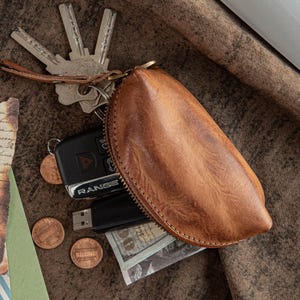 Puede incluir: Un estuche de llaves de cuero marrón con cierre de cremallera, que contiene llaves, un mando de coche, monedas, una unidad USB y algo de efectivo. El estuche de llaves tiene una correa de muñeca y una superficie texturizada.