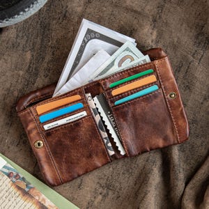 May include: Open brown leather wallet filled with cash, credit cards, and other cards. The wallet has multiple card slots and a zippered compartment. The wallet is open, displaying its contents. The leather has a distressed look.