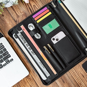 Może przedstawiać: Czarny skórzany portfel z wieloma przegródkami, w tym miejscami na karty, kieszenią na telefon i uchwytami na długopisy. Portfel zawiera tablet, telefon, długopisy i kabel ładujący. Tekst "making us all fat?" jest widoczny na tablecie. Widoczne są również słowa "global trade".