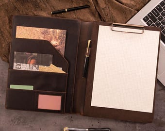 May include: Open brown leather portfolio with a clipboard, pen, and pockets for cards and documents. A laptop and additional pen are visible in the background. The portfolio is open, revealing a blank sheet of grid paper.