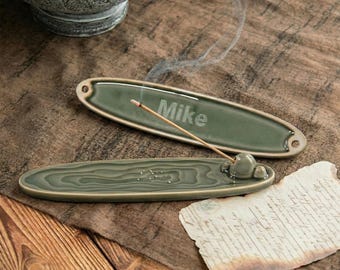 May include: Two green ceramic incense holders with a matching design. One holder has the name "Mike" engraved. The other holder has a small stone-like feature to hold the incense stick. A lit incense stick is in the holder, with smoke rising.