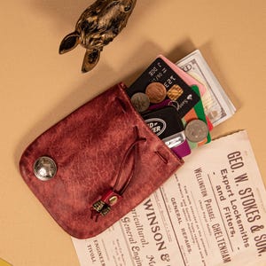 May include: A burgundy leather pouch with a drawstring closure, holding credit cards, coins, and cash. The pouch features a decorative silver button and a small metal accent on the drawstring. A vintage advertisement is partially visible.