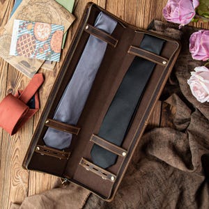 May include: A brown leather tie case with a zipper, holding a gray and a black tie. The case has leather straps to secure the ties. The background includes a wooden surface, a rolled-up tie, and a floral arrangement.
