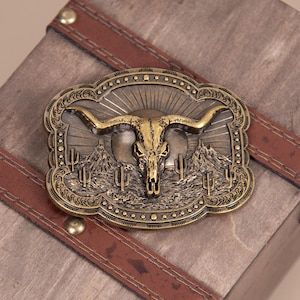 May include: An antique brass-colored belt buckle with a detailed design. The buckle features a bull skull with long horns, mountains, cacti, and a sunburst pattern. The buckle is rectangular with an ornate border.