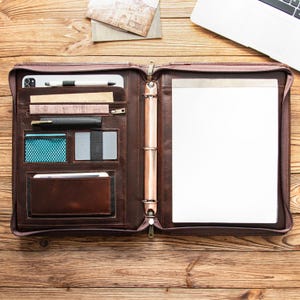 Puede incluir: Portafolio de cuero marrón abierto con múltiples bolsillos, un portalápices y un bloc de notas. El portafolio está abierto, revelando varios compartimentos para tarjetas, un teléfono y otros accesorios. Una computadora portátil y artículos de papelería son visibles en la superficie de madera.