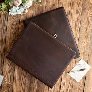 May include: Two dark brown leather portfolios are displayed on a wooden surface. The portfolios have a rectangular shape with rounded corners. A pen and a postcard are next to the portfolios. The portfolios are likely for business or office use.