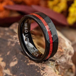 Puede incluir: Anillo de tungsteno negro con un diseño facetado y una incrustación roja vibrante. El anillo está grabado con "W&Q Lydia 4, 2023" en el interior. El anillo se muestra sobre una superficie de piedra texturizada.
