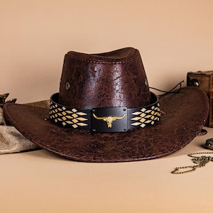 May include: A brown cowboy hat with a textured finish. The hat features a black band with a gold bull head emblem and a geometric pattern. The hat's brim is wide, and the crown is indented. A classic Western-style accessory.