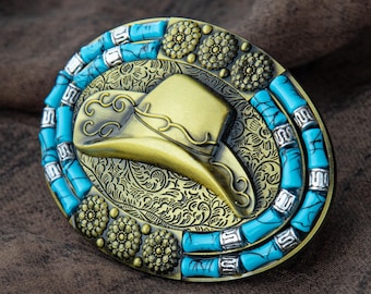 May include: An oval, antique gold-coloured belt buckle with a cowboy hat design. It features turquoise-coloured stones and floral detailing. The hat is set against a detailed, engraved background.
