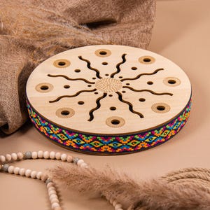 Può includere: Strumento musicale rotondo in legno chiaro, ornato da una fascia colorata. La parte superiore presenta un motivo a forma di sole con linee ondulate e ritagli circolari. Una collana di perline e una pianta secca sono in primo piano.