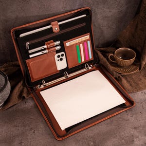 May include: Open brown leather binder with multiple compartments, including pen holders, card slots, and a phone pocket. The binder contains a notepad and a tablet. A cup of coffee sits in the background.