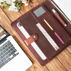 May include: A brown leather portfolio with multiple pockets, a pen holder, and a snap closure. The portfolio is open, revealing a white tablet and various colored cards. A laptop is visible in the background, suggesting a workspace.