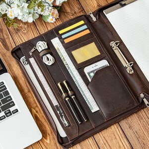 May include: A brown leather portfolio with multiple compartments, including card slots, a phone pocket, and pen holders. The portfolio is open, revealing a notepad, pens, a phone, and a laptop. The background is a wooden surface.