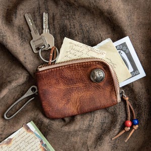 May include: A brown leather key wallet with a zipper, a silver button, and a leather pull with red and blue beads. The wallet is open, revealing keys, a metal clip, and a few pieces of paper, including a dollar bill.