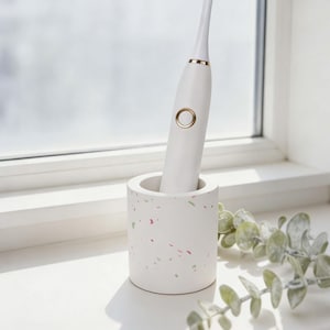 May include: A white electric toothbrush with gold accents is placed in a white speckled holder. The holder has flecks of red and green. The toothbrush is on a windowsill with a sprig of greenery.