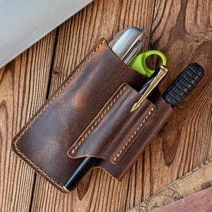 Op de afbeelding: Een bruine leren pennenhouder met een gestikt ontwerp, met een pen, schaar en een multitool. De pen heeft een goudkleurige penpunt. De houder staat op een houten oppervlak.