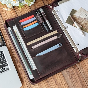 Può includere: Una cartella in pelle marrone con più tasche, contenente un tablet, penne, carte e uno smartphone. La cartella è aperta, rivelando un taccuino e una cartolina vintage. La cerniera è chiusa.