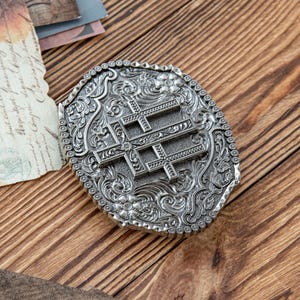 May include: Oval-shaped silver belt buckle with an ornate floral and scroll design. The buckle features three crosses in the center. The border is lined with small, decorative studs. The buckle is set against a wooden background.