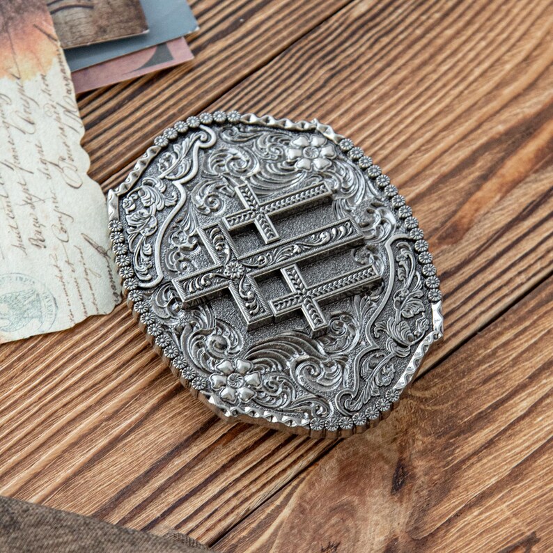 May include: Oval-shaped silver belt buckle with an ornate floral and scroll design. The buckle features three crosses in the center. The border is lined with small, decorative studs. The buckle is set against a wooden background.