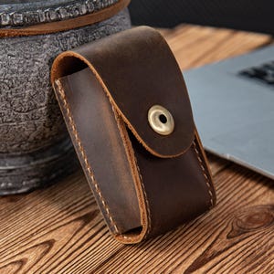 Könnte beinhalten: Eine braune Ledertasche mit Klappe und Messingknopfverschluss. Die Tasche ist rechteckig mit sichtbaren Nähten und einem abgenutzten Aussehen. Die Tasche liegt auf einer Holzoberfläche.