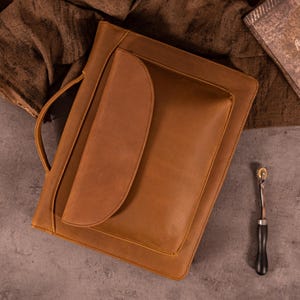 Könnte beinhalten: Eine hellbraune Ledermappe mit Griff und großer Vordertasche. Die Mappe ist rechteckig mit abgerundeten Ecken und einer Klappe, die die Tasche bedeckt. Ein kleines Metallwerkzeug mit Holzgriff liegt auf der Oberfläche.