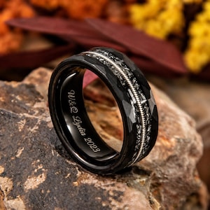 May include: A black ring with a faceted design and a silver string inlay. The ring has the inscription "W&C Lydia 2023" on the inside. The ring is set against a natural stone background.