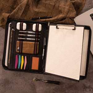 Pode incluir: Pasta de couro preta com vários compartimentos, incluindo ranhuras para canetas, um telefone e um bloco de notas. A pasta também contém um tablet, um par de fones de ouvido brancos e notas autocolantes coloridas. Uma caneta preta e um laptop também são visíveis.