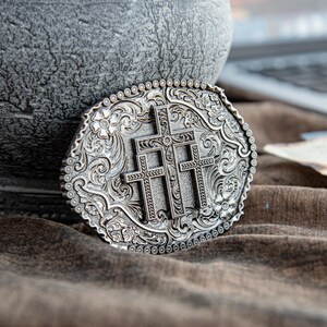 May include: Silver-toned oval belt buckle featuring three crosses at the center, surrounded by floral and scrollwork designs. The buckle has a detailed, textured surface and a beaded border. A religious-themed accessory.