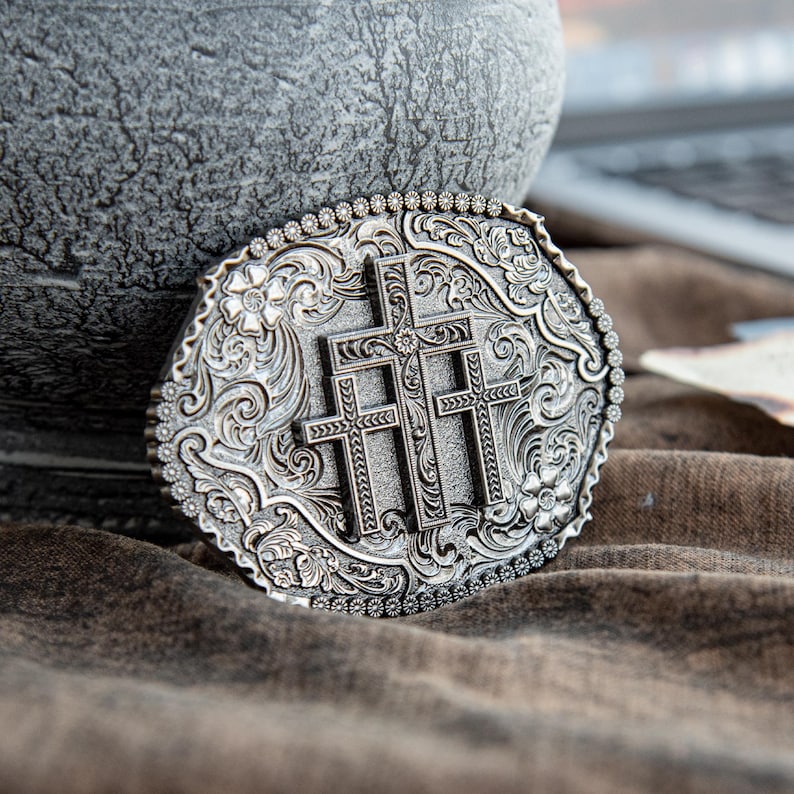 May include: Silver-toned oval belt buckle featuring three crosses at the center, surrounded by floral and scrollwork designs. The buckle has a detailed, textured surface and a beaded border. A religious-themed accessory.
