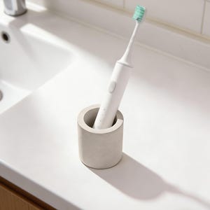 Puede incluir: Un cepillo de dientes eléctrico blanco con un cabezal verde se coloca en un pequeño portacepillos cilíndrico de hormigón gris claro. El soporte está sobre una encimera blanca junto a un lavabo.