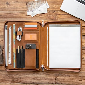 May include: Open brown leather portfolio with multiple compartments, including card slots, pen holders, and a phone pocket. A notepad, tablet, and charging cables are visible. The portfolio is open on a wooden surface, with a laptop in the background.