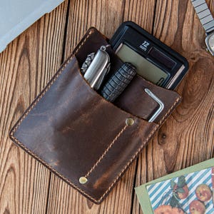 May include: A brown leather pocket organizer with a calculator, multi-tool, and other small tools. The organizer is open, revealing the contents. The background is a wooden surface.