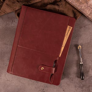 May include: A burgundy leather portfolio with a snap closure and a pocket holding a vintage ticket. A rotary wheel tool with a wooden handle rests nearby. The portfolio is on a gray surface.
