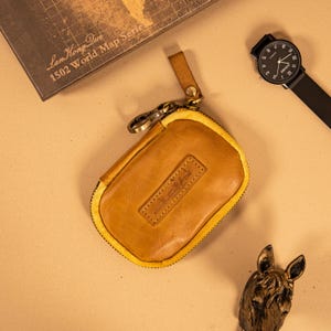 May include: A tan leather key case with a yellow zipper and a small leather strap. A black watch with a black band and a bronze horse head figurine are also visible. The key case has a stitched leather patch.