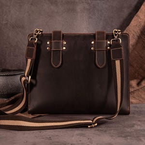 Könnte beinhalten: Dunkelbraune Leder-Umhängetasche mit rechteckiger Form. Die Tasche hat einen langen, verstellbaren Schulterriemen mit einem gestreiften Muster in Beige und Braun. Die Tasche hat zwei Lederriemen mit Messingnieten und Beschlägen.