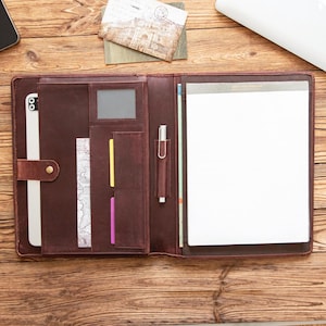 May include: Open, brown leather portfolio with multiple pockets, a pen holder, and a notepad. The portfolio is open, revealing a white notepad and a tablet. A postcard and a laptop are visible in the background.