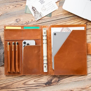 May include: Open, brown leather organizer with multiple pockets for cards, pens, and a smartphone. A silver tablet is visible in a large pocket. The organizer is open on a wooden surface.
