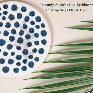 May include: White round coaster with a pattern of blue watercolor blueberries. The coaster has the text "Instantly Absorbs Cup Residue Desktop Stays Dry & Clean". Green palm leaves are in the background.