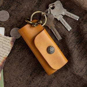 Pode incluir: Um porta-chaves de couro castanho claro com fecho de botão de latão e um porta-chaves de latão. Várias chaves estão presas ao anel. O porta-chaves é retangular com uma aba curva. O fundo é uma superfície castanha texturizada.