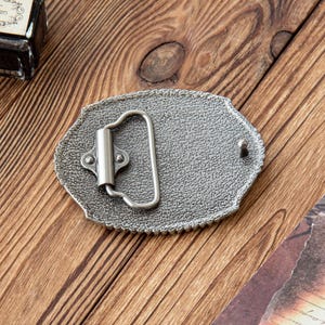May include: A silver-colored metal belt buckle with an oval shape and a textured surface. The buckle has a decorative edge and a hinged bar for the belt. The buckle is on a wooden surface.