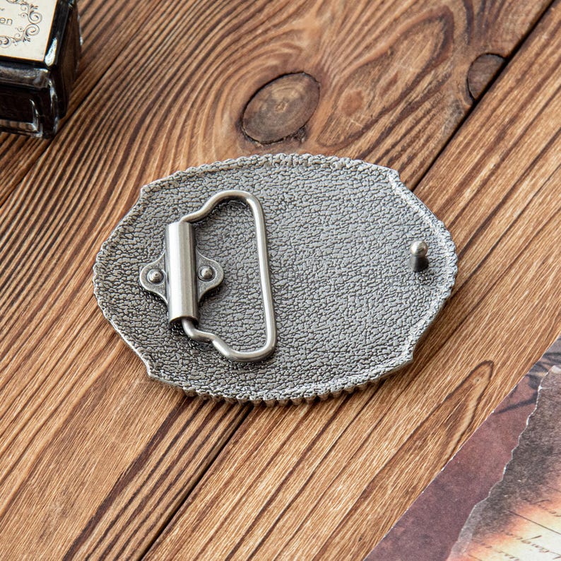 May include: A silver-colored metal belt buckle with an oval shape and a textured surface. The buckle has a decorative edge and a hinged bar for the belt. The buckle is on a wooden surface.