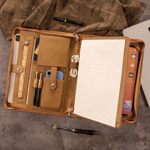 Peut inclure: Une mallette en cuir beige ouverte, révélant un bloc-notes, une tablette et un support pour téléphone. La mallette contient des stylos, un passeport et un câble de chargement. Un ordinateur portable est partiellement visible en arrière-plan.